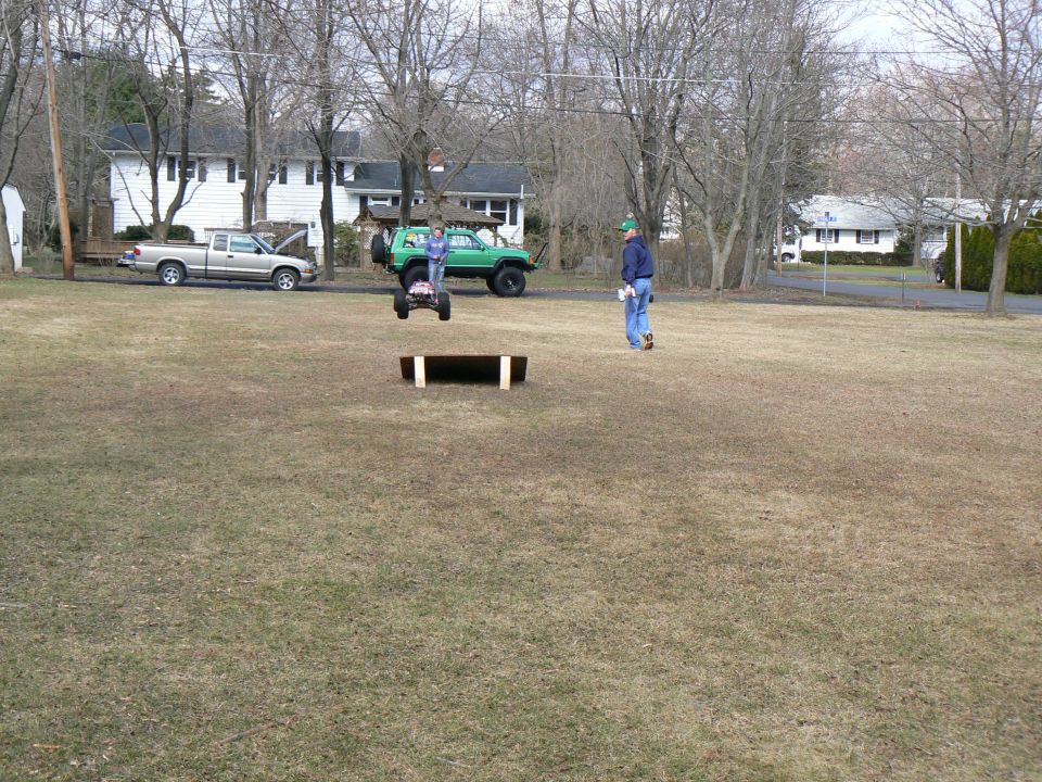 Remote control car mid-air over a ramp, with a green SUV and a pickup truck in the background