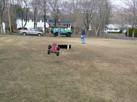 1994 Ford Bronco - A red remote control monster truck is captured mid-air as it jumps over a ramp in a grassy yard.