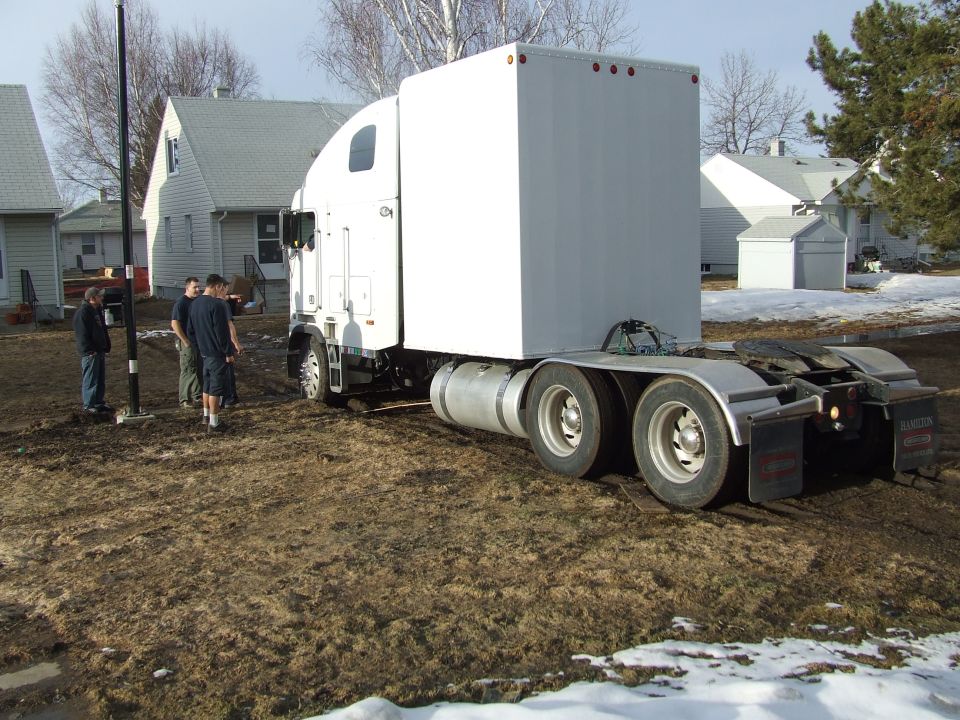 White semi-truck tractor with a white box trailer parked on muddy ground near houses
