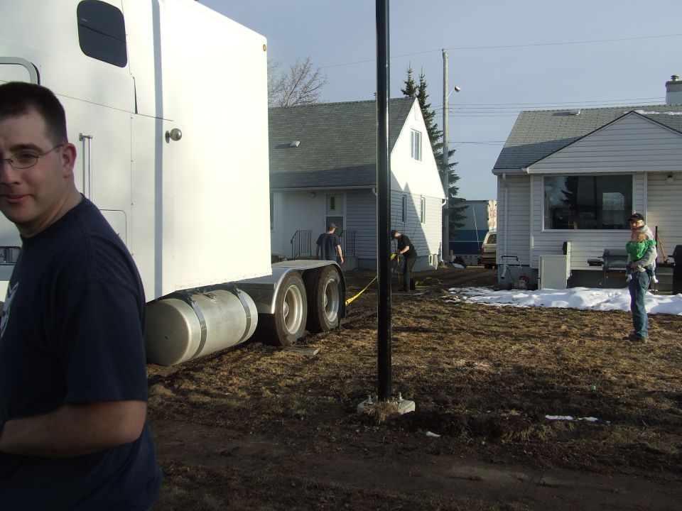 man in foreground, semi-truck trailer, people working in yard, houses in background