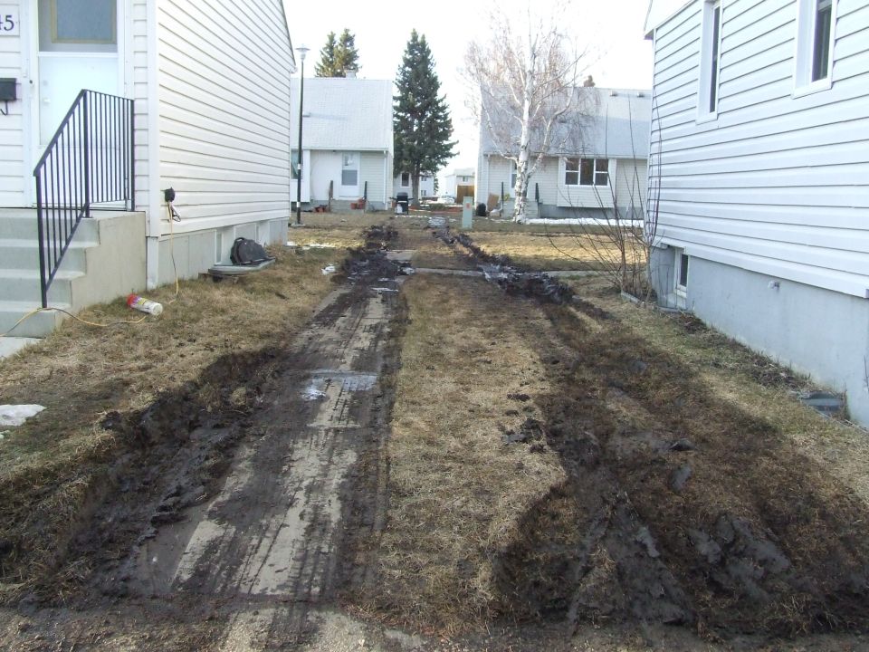 Muddy driveway between two houses with tire tracks and disturbed grass