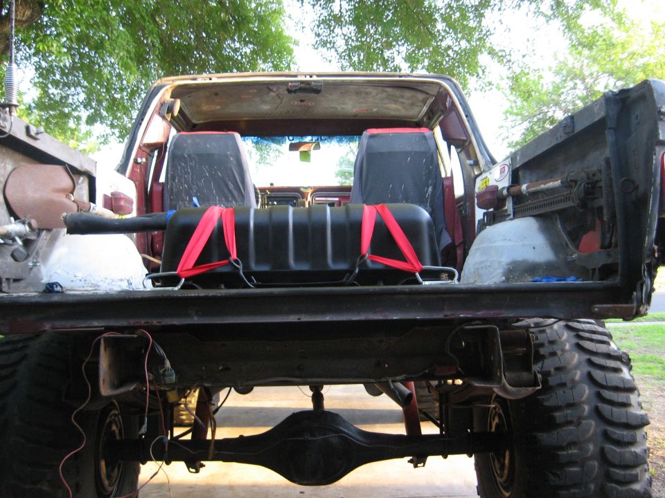 Rear view of a red 1978 Ford Bronco with its tailgate down, showing a black auxiliary fuel tank secured with red straps inside