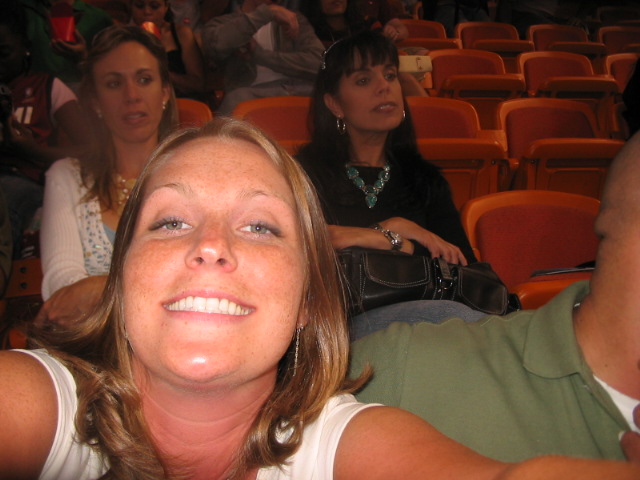 Woman with freckles smiles at camera in stadium seating, with other people behind her