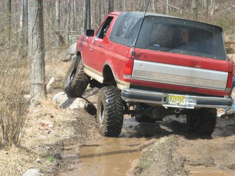 1990 Ford Bronco - This red SUV is shown off-roading on a rocky and muddy path through a wooded area.