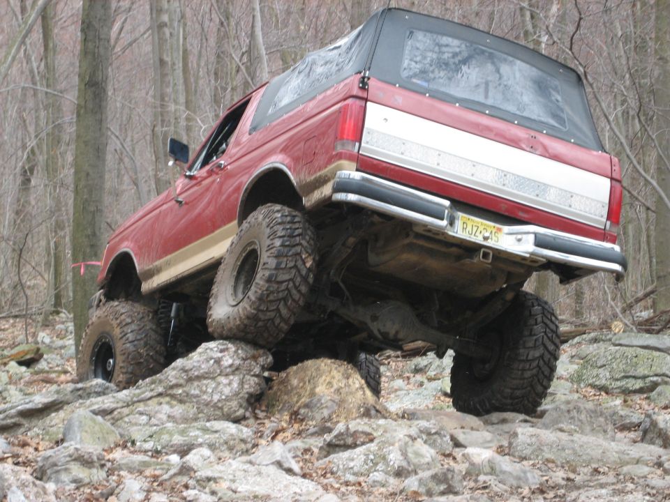 Red 1990 Ford Bronco with a soft top, lifted suspension, and large off-road tires, climbing over rocks