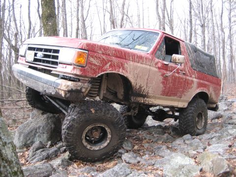 1990 Ford Bronco - A red Ford Bronco navigates a rocky off-road trail, its front-left tire high in the air.