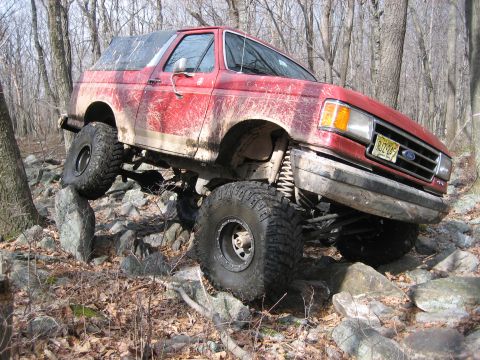 1990 Ford Bronco - Red 1990 Ford Bronco tackles a challenging off-road course with one wheel lifted.