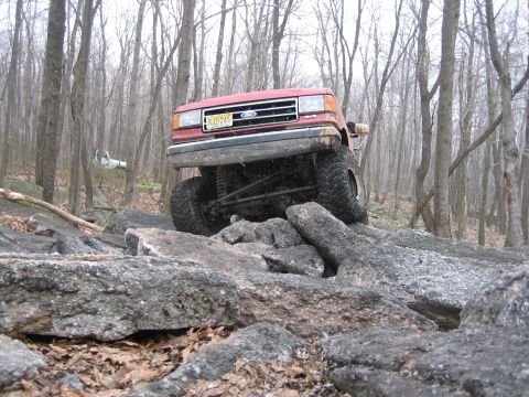 1990 Ford Bronco - A red Ford Bronco navigates a rocky trail in a wooded area.