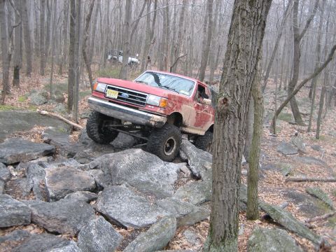 1990 Ford Bronco - A red 1990 Ford Bronco tackles a challenging rocky trail in a wooded area, with another vehicle visible in the background.