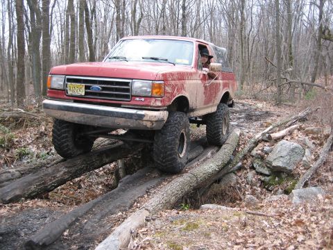 1990 Ford Bronco - This red Ford Bronco is navigating a challenging off-road course made of logs and rocks in a forest.