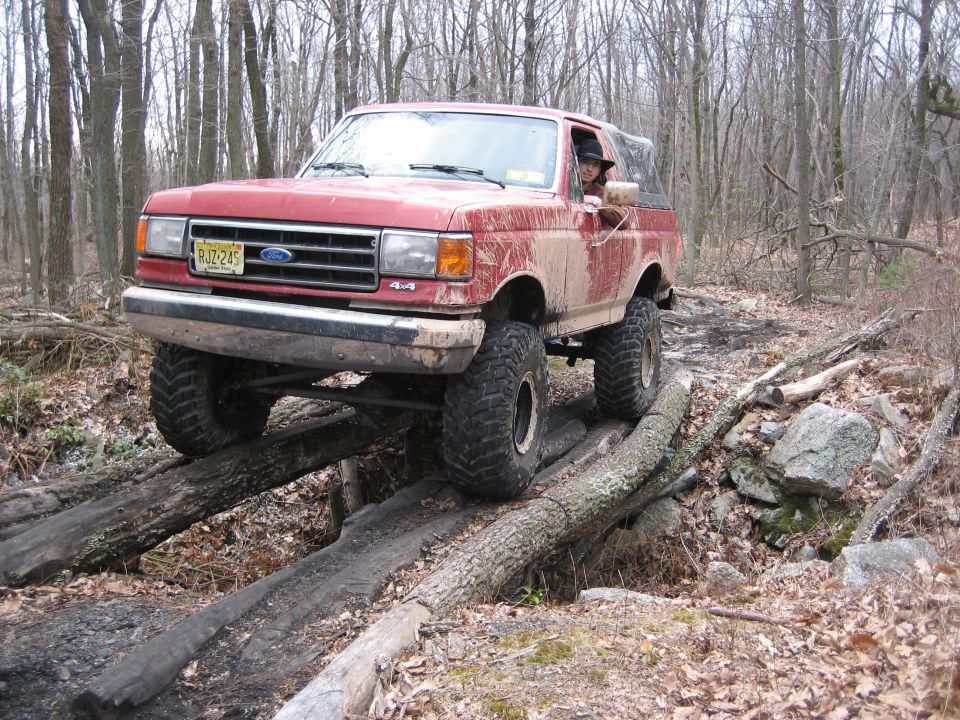 Red Ford Bronco driving over logs on a wooded trail. Driver wearing a hat is visible