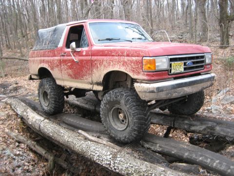 1990 Ford Bronco - A red 1990 Ford Bronco navigates a challenging off-road course on a wooded trail.