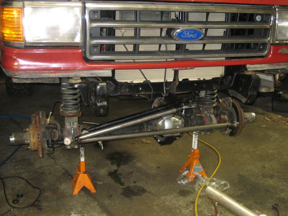 Red Ford truck front end with grille, headlights, and exposed axle on jack stands