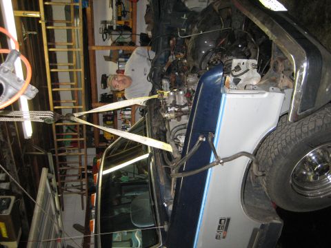 1990 Ford Bronco - A mechanic works on the engine of a Ford F-150 in a garage, with straps holding the engine in place.