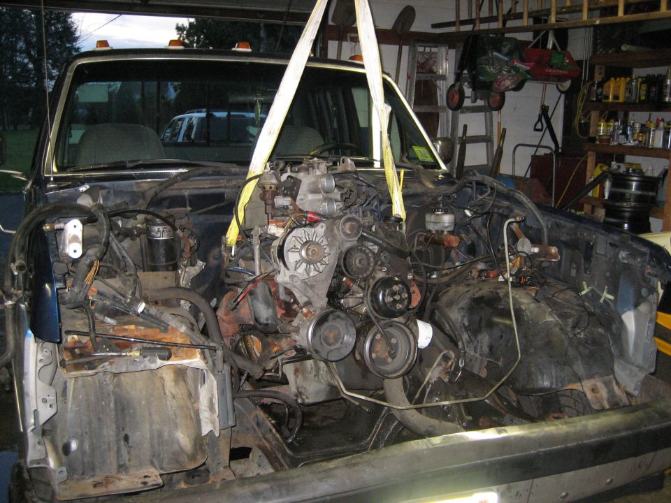 Engine being lifted out of a dark blue truck with a hoist. The front end of the truck is disassembled