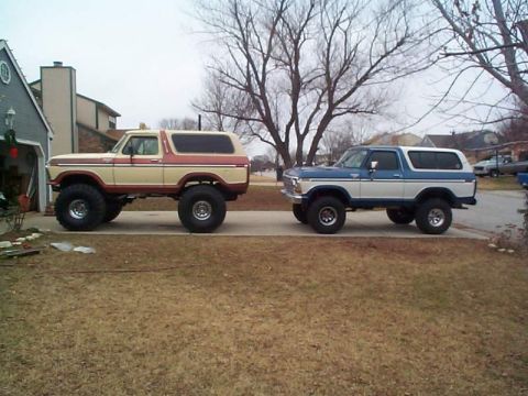1978 Ford Bronco - Two lifted full-size Ford Broncos parked side by side in a suburban driveway, shot in profile, one in tan over red two-tone, the other in b…