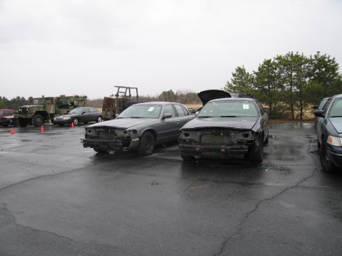 1991 Ford LTD Crown Victoria - Two damaged sedans are parked in an outdoor lot, with military vehicles in the background.