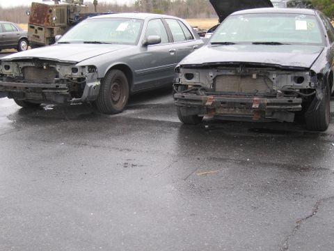 1991 Ford LTD Crown Victoria - Two wrecked 1991 Ford Crown Victoria LTDs sit side-by-side in a junkyard, their front ends heavily damaged.