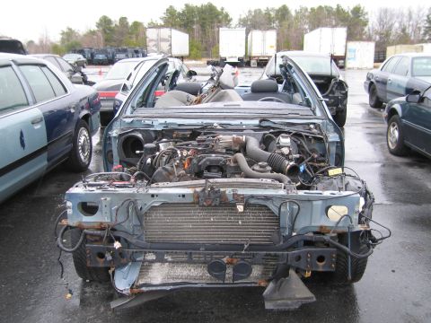 1991 Ford LTD Crown Victoria - A heavily damaged sedan sits in a junkyard, its front end dismantled and engine exposed to the elements.