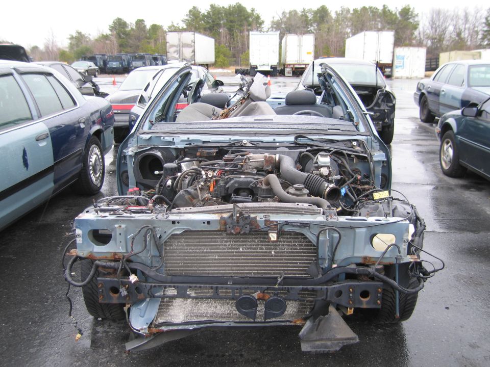 Wrecked light blue sedan with front end removed, engine exposed, parked in a lot with other vehicles and trailers