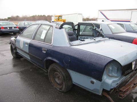 1991 Ford LTD Crown Victoria - A heavily damaged police car sits in a lot, missing its roof and several body panels, with other vehicles visible in the background.