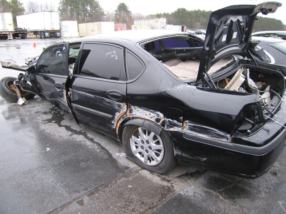 Wrecked black sedan with doors open, flat tires, and damaged front end. Trunk is open, showing interior damage