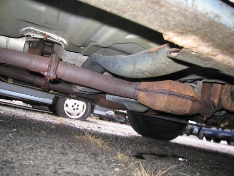1991 Ford LTD Crown Victoria - The undercarriage of a vehicle reveals a rusty exhaust system and a flat tire, with other vehicles visible in the background.