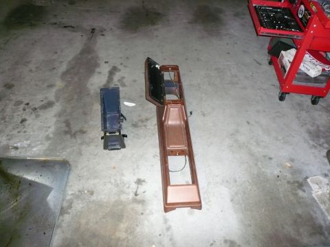 1975 Ford Ranchero - Two car interior console parts, one brown and one black, are laid out on a concrete floor next to a red toolbox.