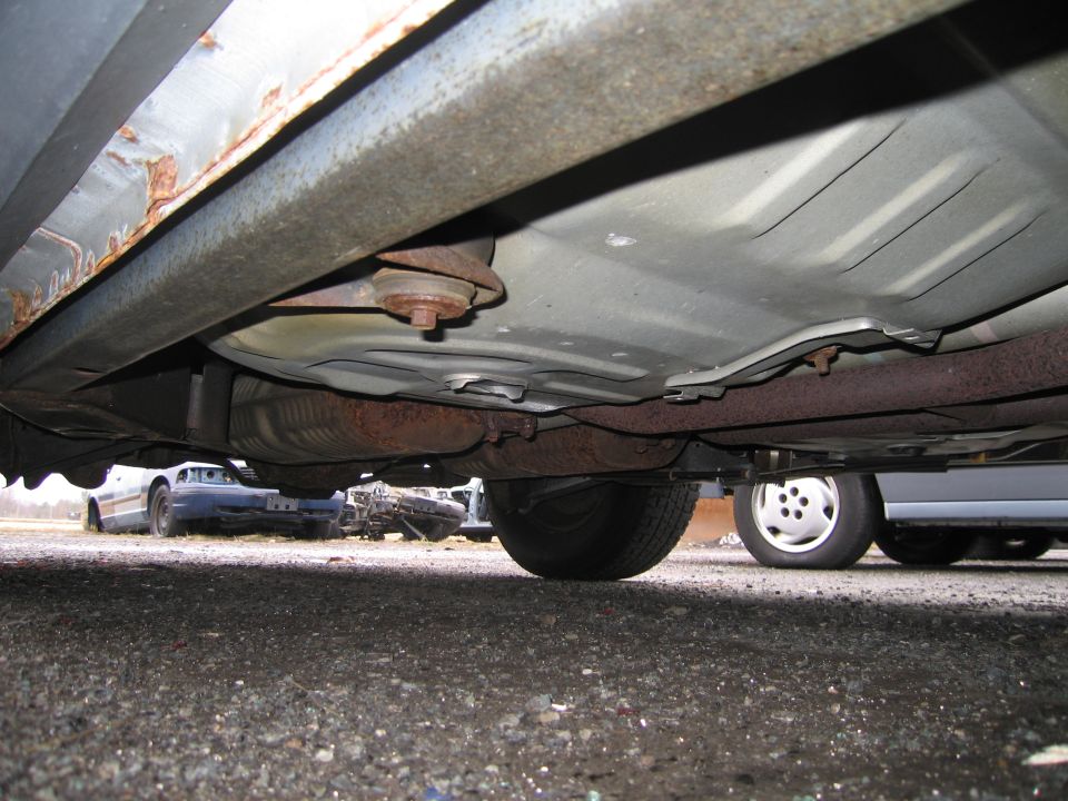 Underside view of a car showing rust on the frame and exhaust components. Other vehicles are visible in the background