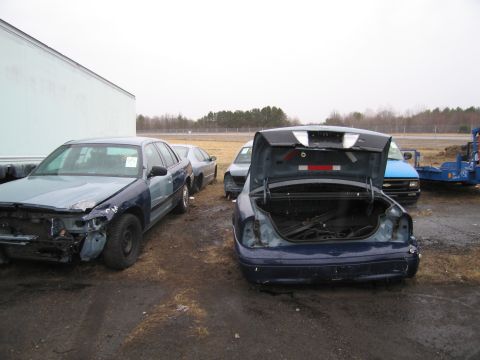 1991 Ford LTD Crown Victoria - A damaged blue 1991 Ford Crown Victoria LTD sits in a junkyard with its trunk open, alongside other vehicles.
