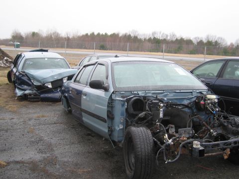 1991 Ford LTD Crown Victoria - Two Ford Crown Victorias, one heavily damaged and the other stripped, sit in a junkyard.