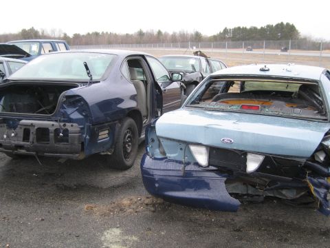 1991 Ford LTD Crown Victoria - Two heavily damaged vehicles are parked on an asphalt lot with trees in the background.