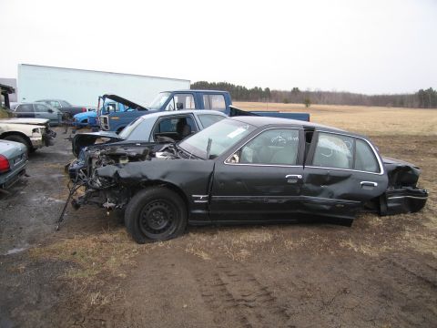1991 Ford LTD Crown Victoria - A damaged dark sedan sits in a junkyard with other vehicles visible in the background.