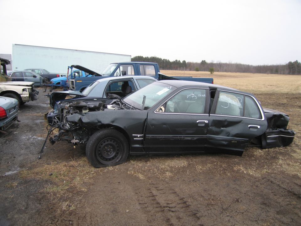 Dark sedan with flat front tire, dented side, and damaged rear, parked in a junkyard