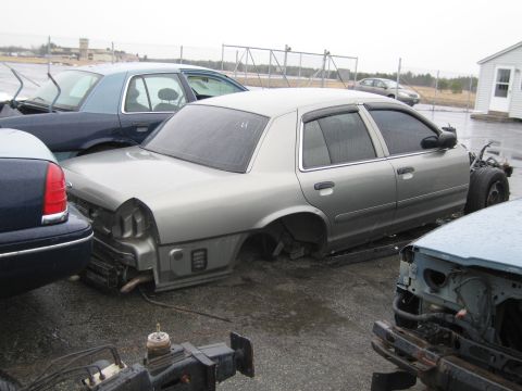 1991 Ford LTD Crown Victoria - A damaged silver sedan sits in a junkyard, stripped of its wheels and front components.