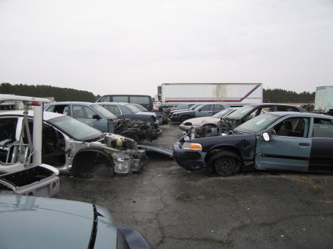 1991 Ford LTD Crown Victoria - A collection of damaged vehicles sits in a junkyard under an overcast sky.