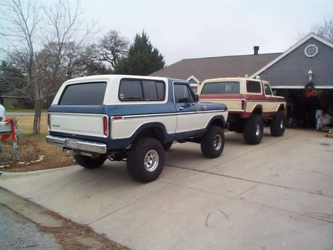 1978 Ford Bronco - Two classic Ford Broncos, modified with lift kits, are parked side-by-side in front of a residential garage.