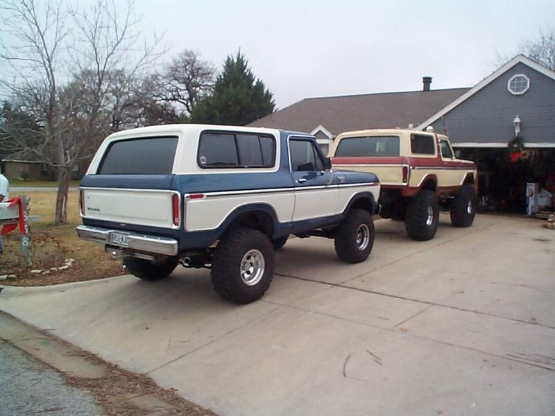 Two lifted Ford Broncos parked on a driveway, one white and blue, the other tan and red