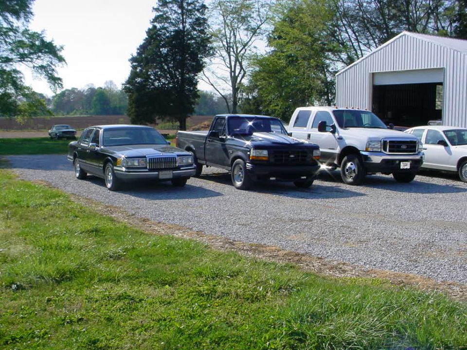 Dark sedan, black pickup truck, white dually pickup truck, and white sedan parked on gravel lot