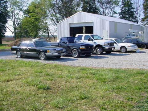 1972 Ford Ranchero - A collection of vehicles are parked on a gravel lot in front of a large metal building on a sunny day.