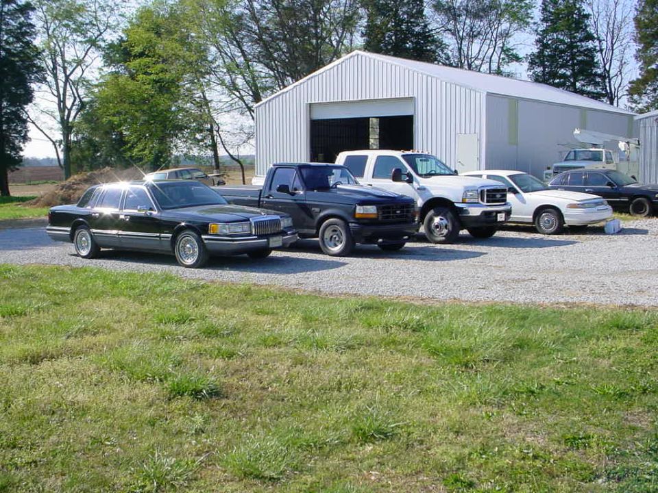 Several vehicles parked in front of a metal barn, including a black sedan, a dark pickup truck, and a white truck