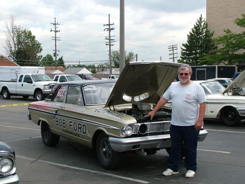 1972 Ford Ranchero - A man stands next to a gold 1964 Ford Falcon, pointing to its open hood and engine. Another car is visible in the background.