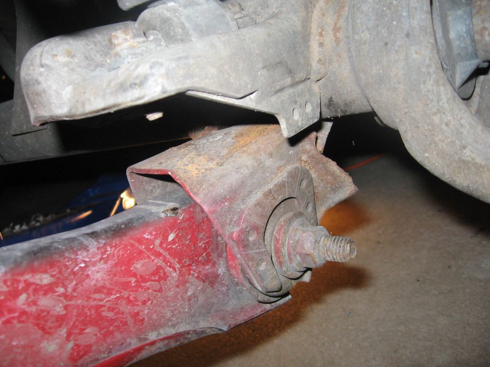Close-up of a rusty red suspension component with a bolt and flange, attached to a larger gray metal assembly