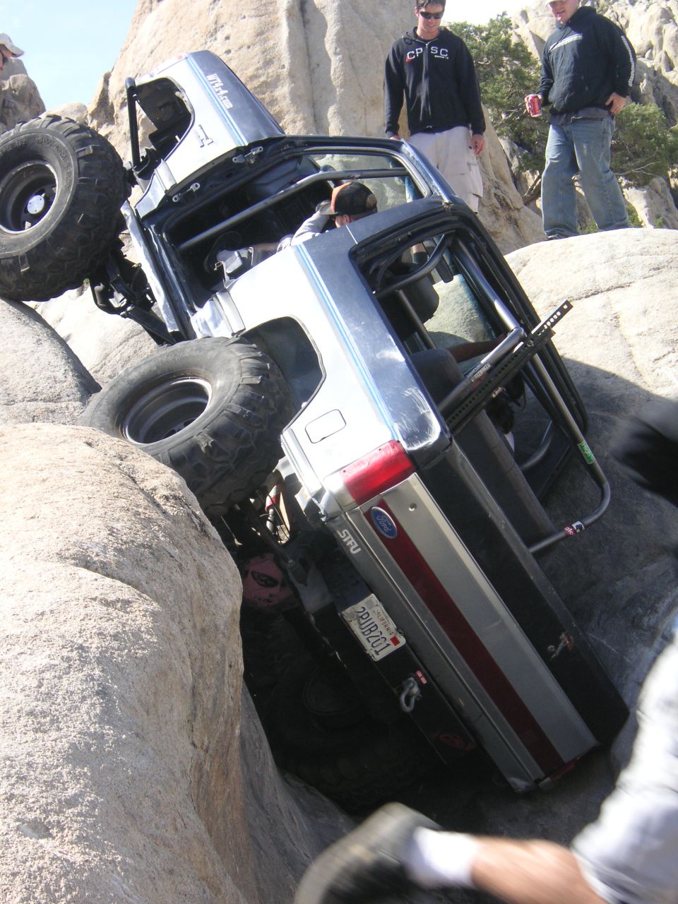 Off-road vehicle tilted precariously between large rocks, with people watching