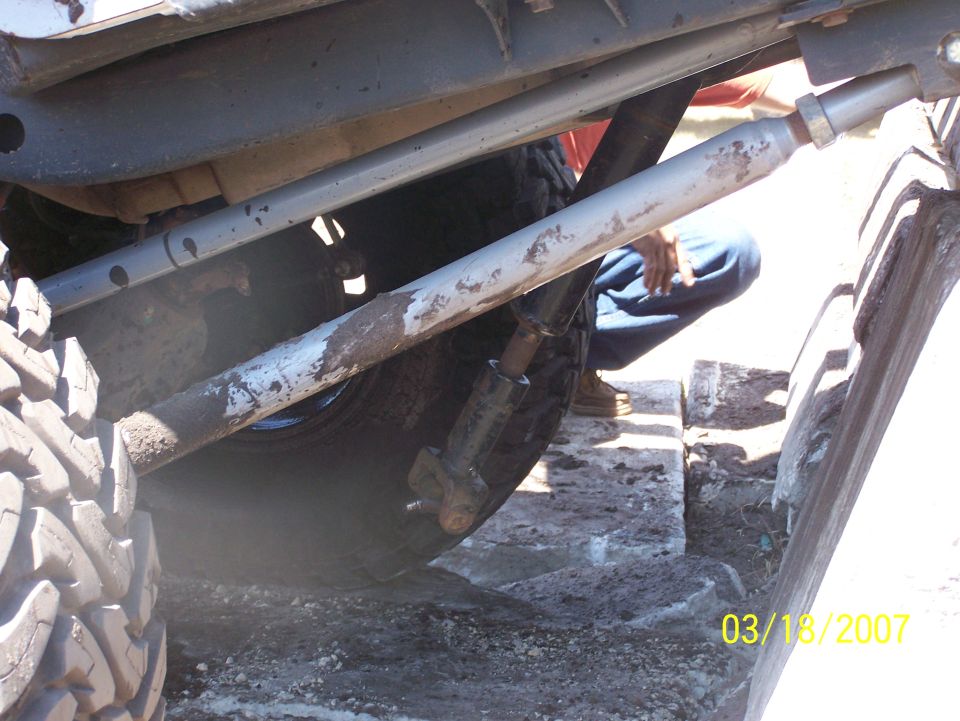 Undercarriage of a vehicle with large tires, showing suspension components and mud. A person is visible in the background