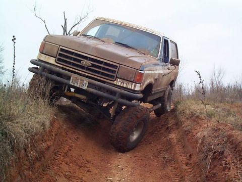 1978 Ford Bronco - Mud-covered brown Ford Bronco with off-road tires, tube bumper and roof rack flexing as it climbs out of a deep rutted dirt trail.