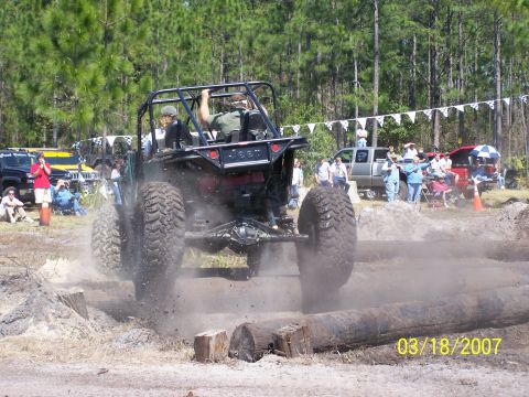 1989 Ford Bronco - An off-road buggy with massive tires powers through a log obstacle course, kicking up dust.
