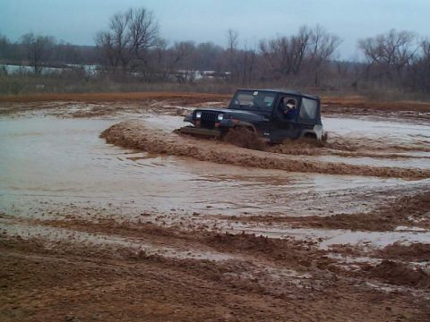 1978 Ford Bronco - A black Jeep Wrangler navigates a muddy track, kicking up dirt and water.