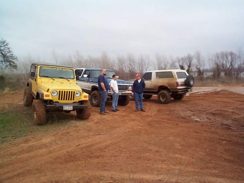 1978 Ford Bronco - Three off-road vehicles and three men stand on a dirt lot under an overcast sky.