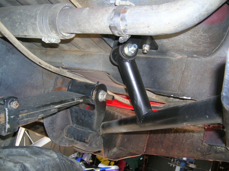 Underbody view of a black tubular link bolted to a frame bracket beside exhaust pipe